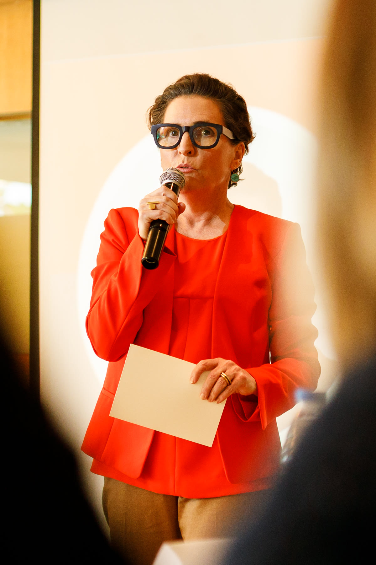A speaker in a red dress addressing the room with a microphone.